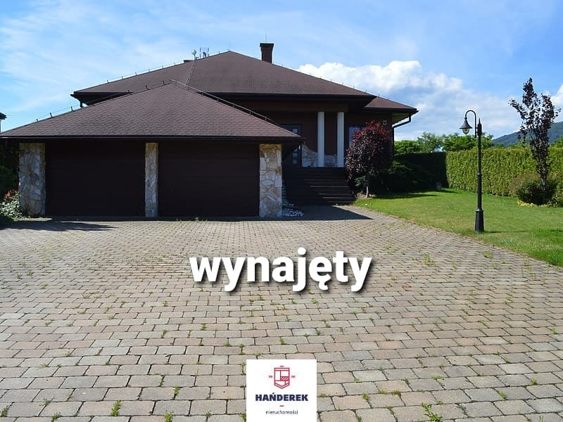 Duży dom jednorodzinny z brązowym dachem i dwustanowiskowym garażem, obok zadbany trawnik i oświetlenie zewnętrzne; na brukowanym podjeździe widoczny napis 'wynajęty' i logo agencji nieruchomości.