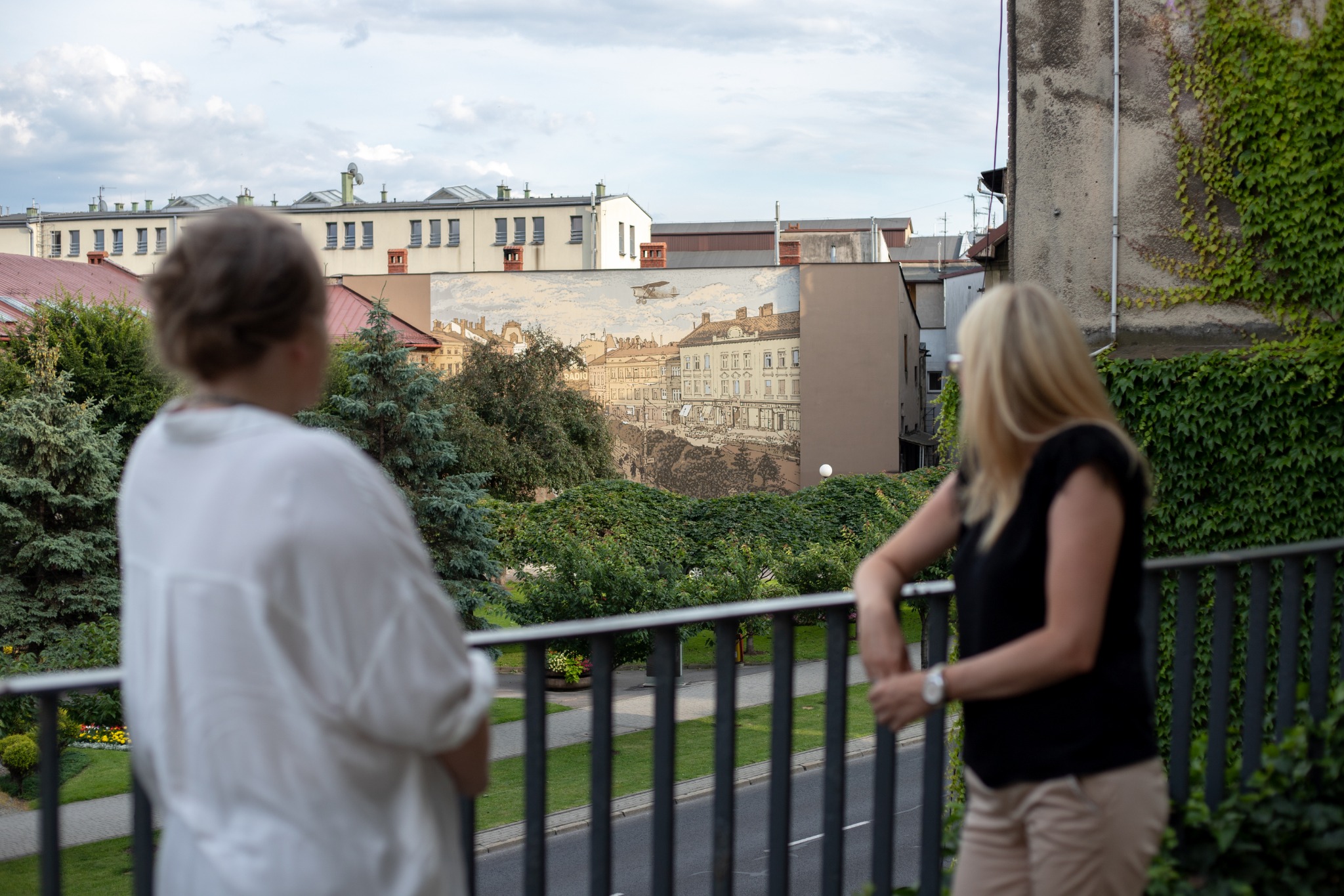 Dwie kobiety stoją na balkonie z metalową balustradą, obserwując mural na budynku naprzeciwko, przedstawiający historyczny widok miasta z lotu ptaka, zieleń drzew i krzewów wypełnia przestrzeń...