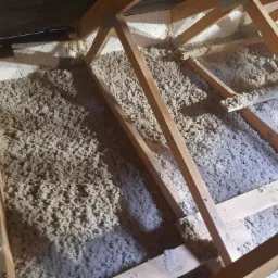 Poddasze z drewnianą konstrukcją wypełnione szarą wełną mineralną, widoczne fragmenty białej piany izolacyjnej na ścianach.