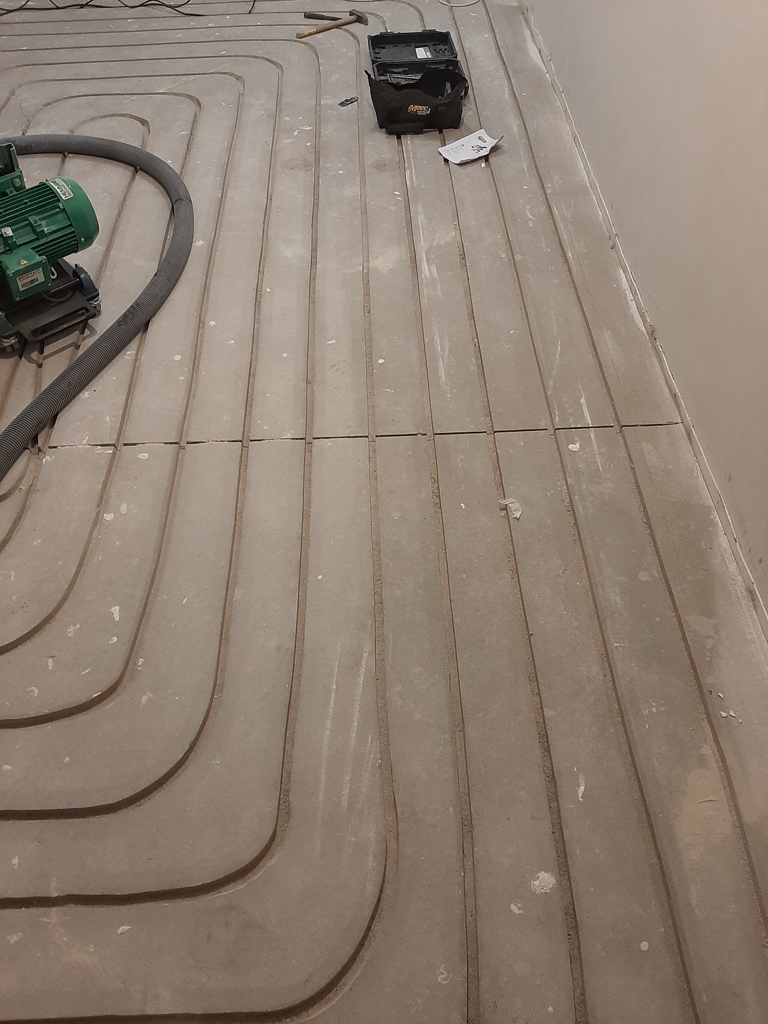 Montaż ogrzewania podłogowego w systemie rowkowym, widoczna frezarka do rowków, narzędzia i dokumentacja na betonowej posadzce.