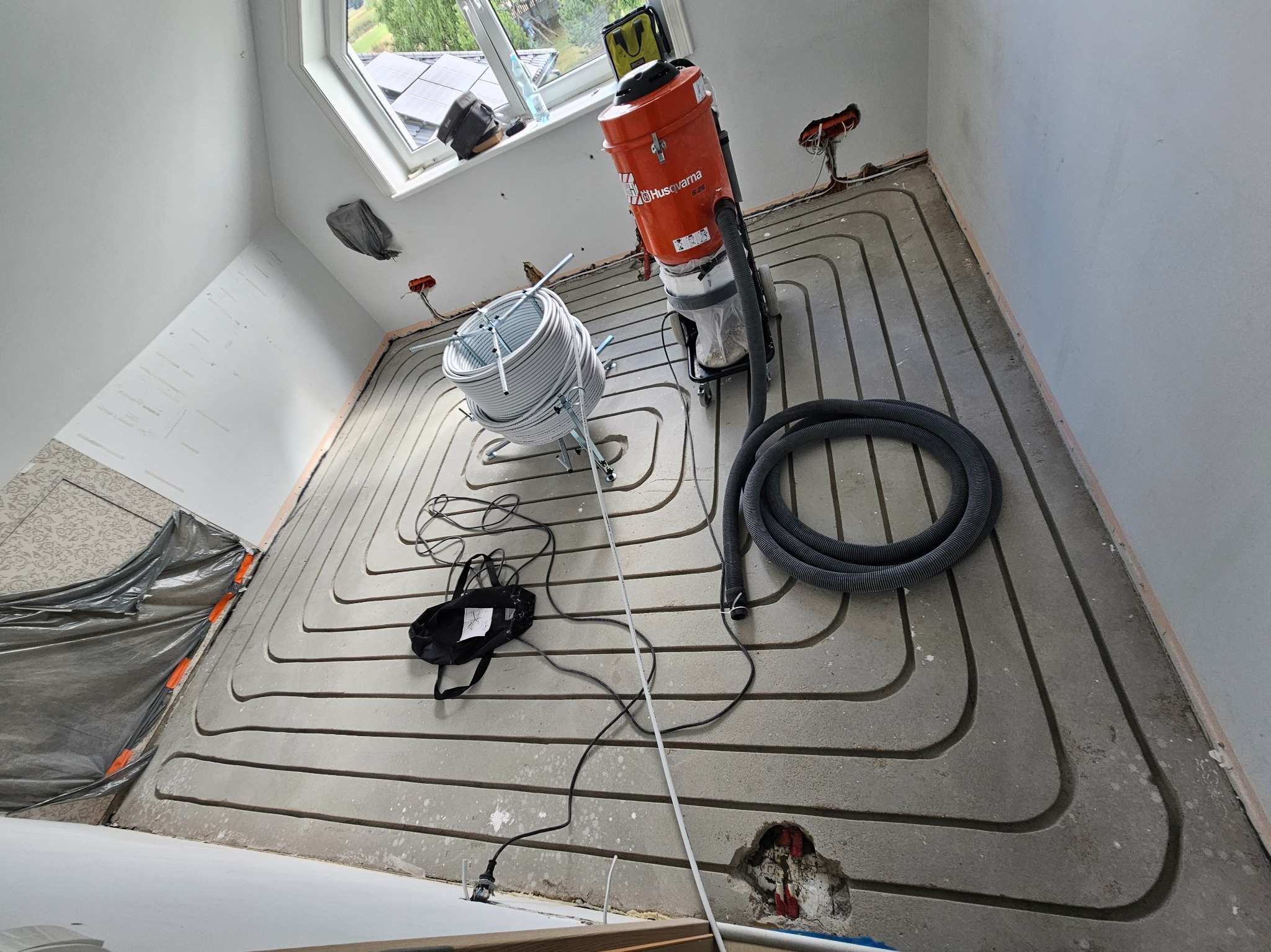 Montaż ogrzewania podłogowego w szarej wylewce. Widoczny zwój rur, odkurzacz przemysłowy Husqvarna i fragment okna dachowego. Instalacja w trakcie realizacji.