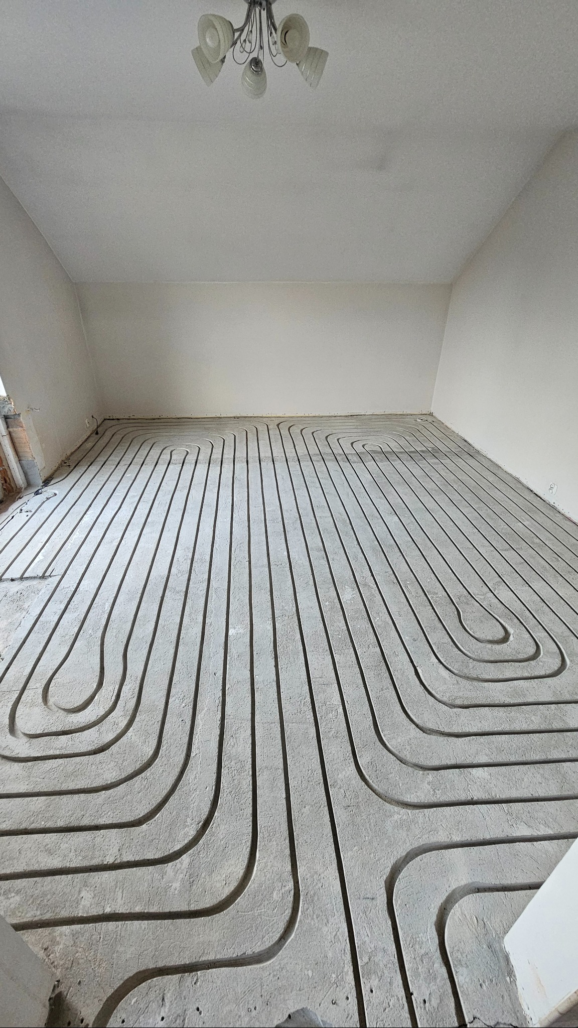 Białe rury ogrzewania podłogowego ułożone w jastrychu, widok z góry na instalację przed zalaniem posadzki. W tle surowy stan ścian i sufitu.