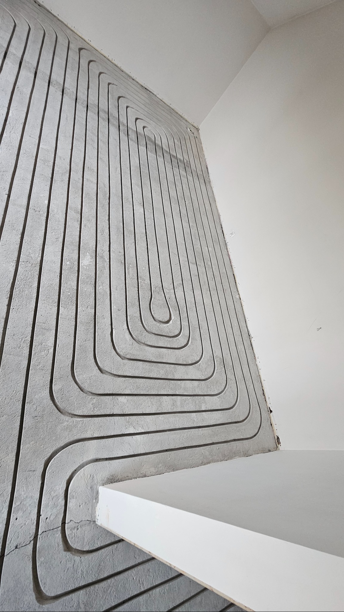 Szare rury ogrzewania podłogowego ułożone w betonowej wylewce, widoczne przed zakryciem posadzką. Biała ściana i fragment podłogi w tle. Ujęcie z dołu.