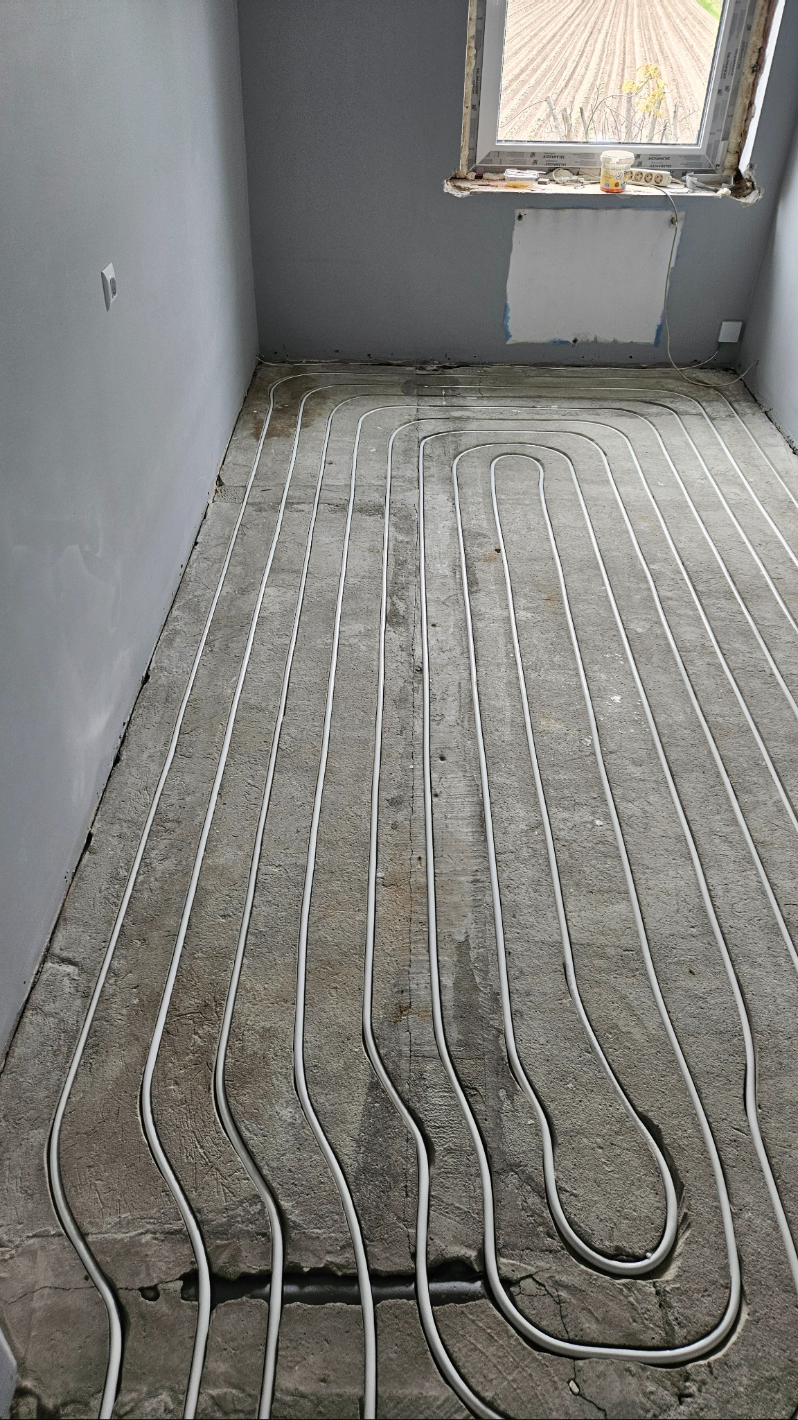 Białe rury ogrzewania podłogowego ułożone na szarym betonie w surowym wnętrzu z oknem. Widok z góry na instalację przed wylaniem posadzki.