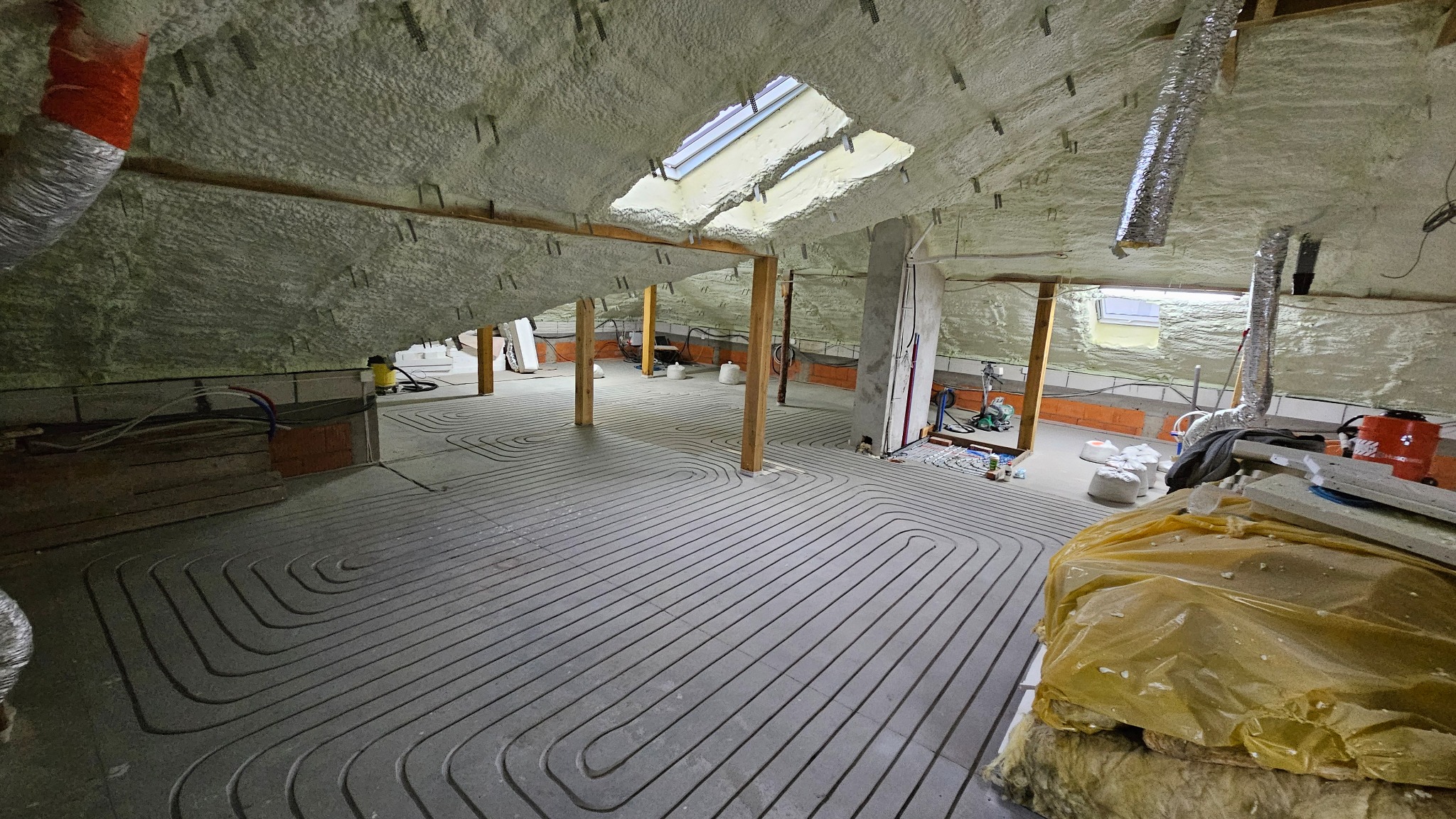 Poddasze z ociepleniem pianą i instalacją ogrzewania podłogowego w szarym betonie. Widoczne drewniane słupy i okno dachowe. Elementy instalacji i narzędzia.