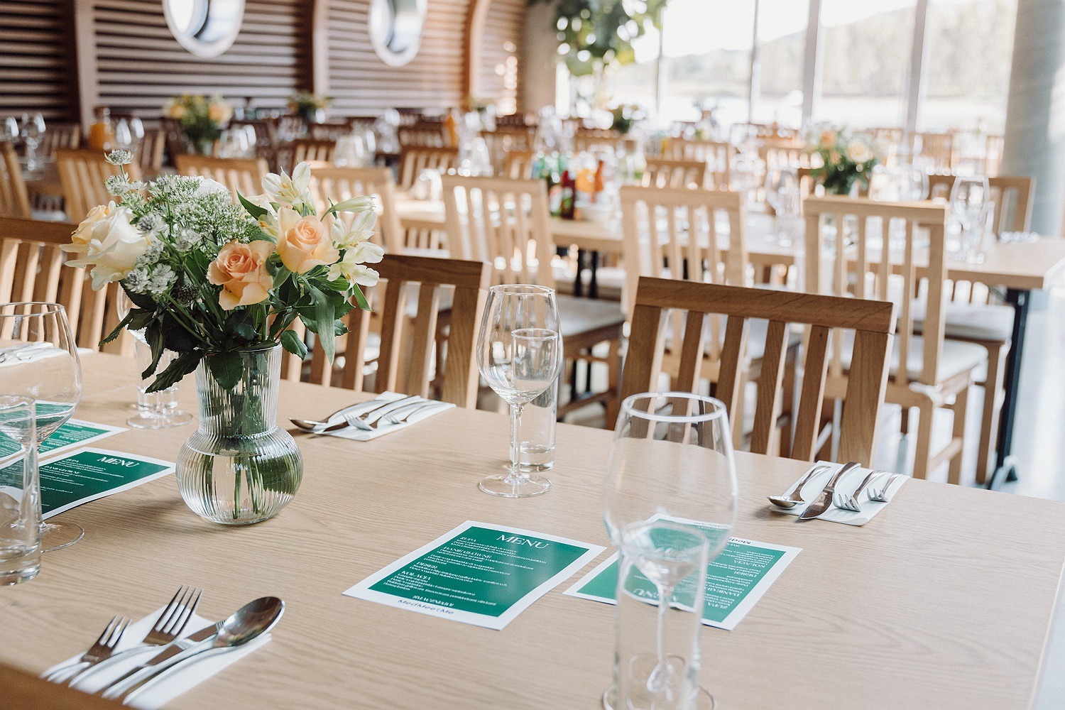 Ustawiony stół z bukietem różowych róż w szklanym wazonie, nakrycia i menu na zielonych kartkach, w tle rzędy krzeseł i stołów w jasnym, przestronnym wnętrzu sali restauracyjnej.