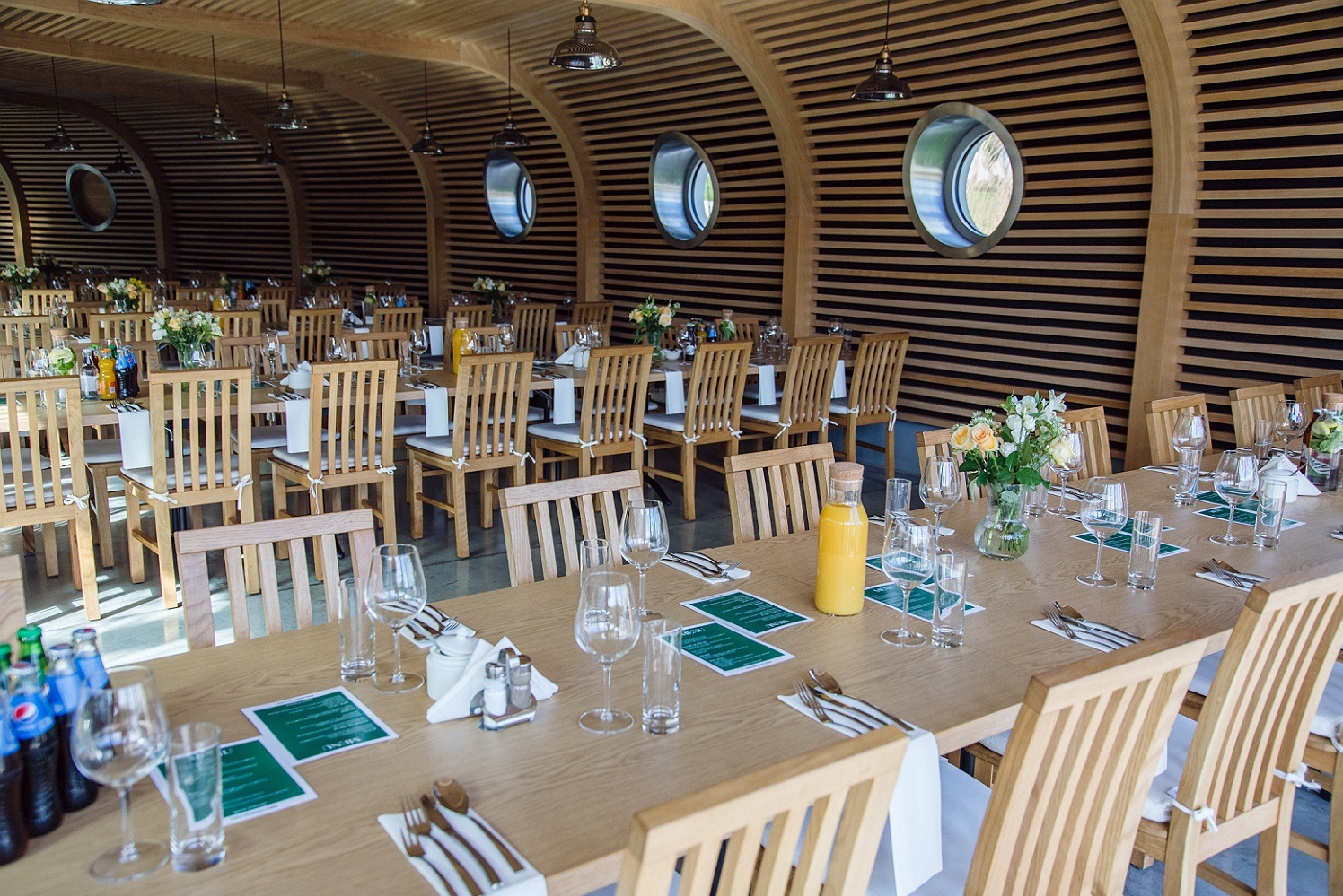 Sala bankietowa z długimi stołami nakrytymi dla gości, drewniane krzesła, dekoracje kwiatowe, napoje i okrągłe okna w tle.