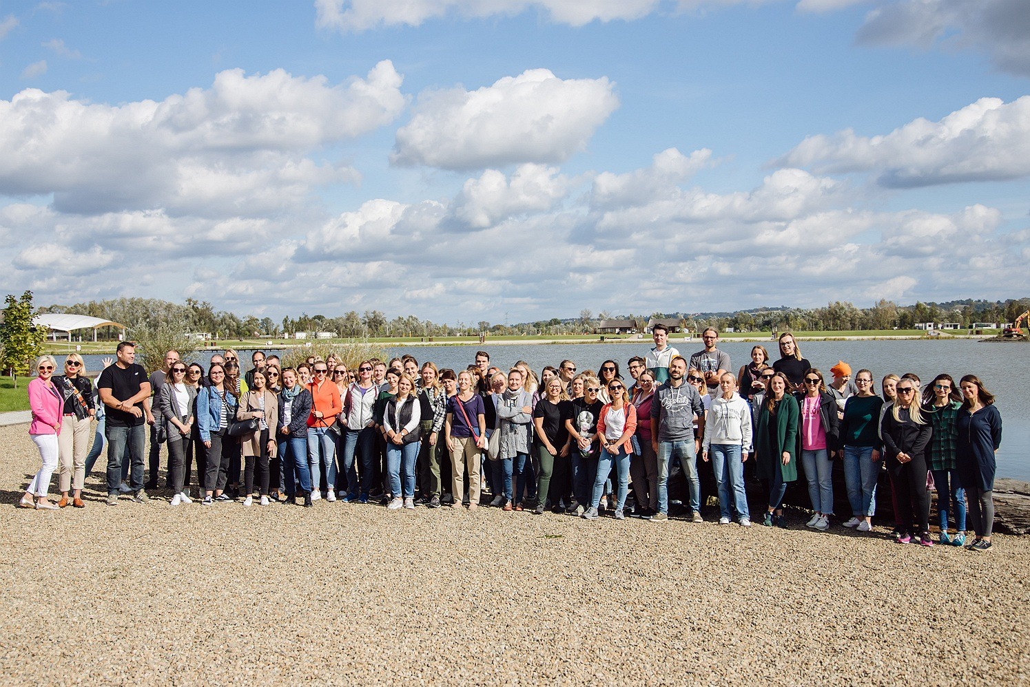 Duża grupa osób pozuje do zdjęcia grupowego na tle jeziora i błękitnego nieba z chmurami, ubrani w codzienne stroje, stoją na żwirowym terenie.