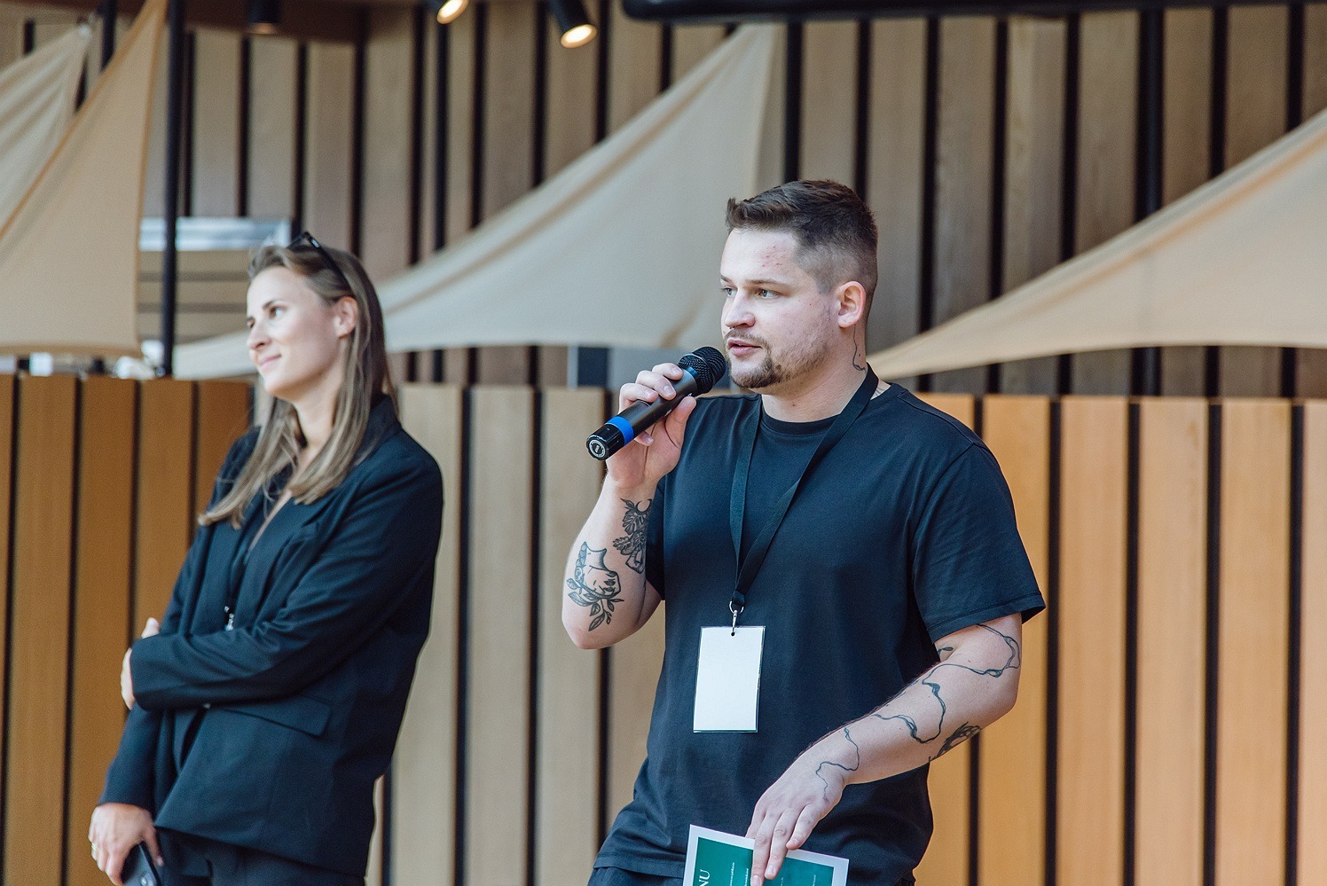 Prelegent z mikrofonem podczas prezentacji na tle drewnianej ściany z dekoracjami żeglarskimi, obok stoi kobieta w czarnym żakiecie.