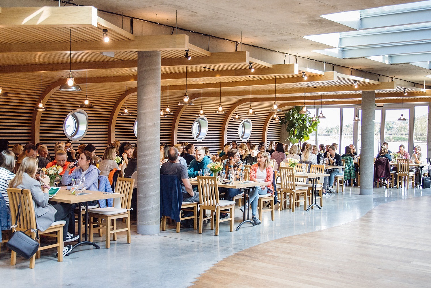 Wnętrze przestronnej sali restauracyjnej w Krakowie, wypełnionej osobami siedzącymi przy drewnianych stołach podczas wydarzenia, z widocznymi dekoracjami kwiatowymi i oknami z widokiem na zewnątrz.