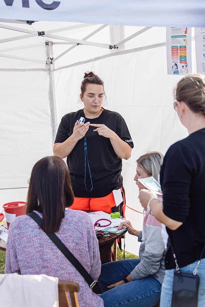 Instruktorka w czarnym uniformie MedAid demonstruje użycie pulsoksymetru uczestnikom pod namiotem podczas szkolenia z pierwszej pomocy.