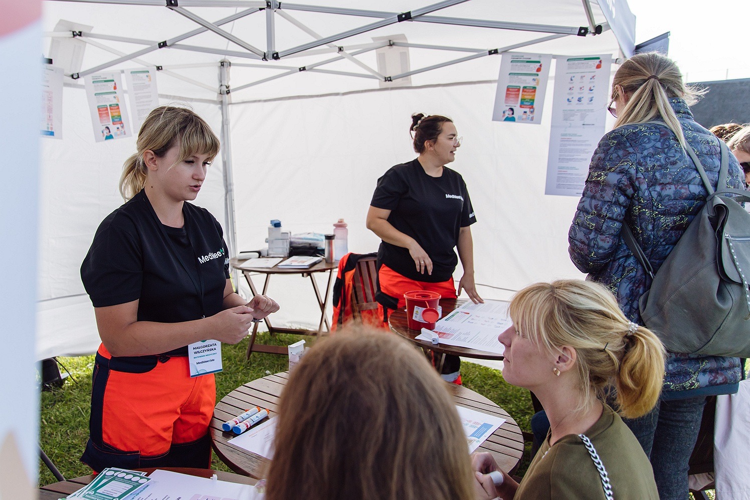 Stanowisko informacyjne kursu pierwszej pomocy pod namiotem, ratownicy w czarnych koszulkach i pomarańczowych spodniach prezentują materiały szkoleniowe zainteresowanym osobom.