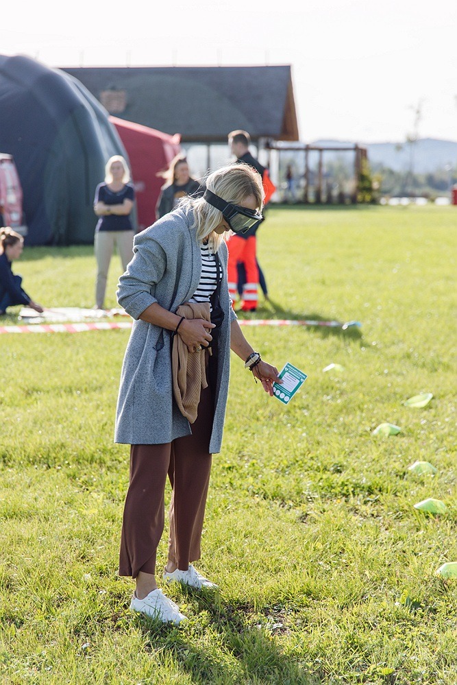 Kobieta z zasłoniętymi oczami trzymająca kartkę na trawniku podczas gry terenowej, w tle grupa osób i duży namiot.