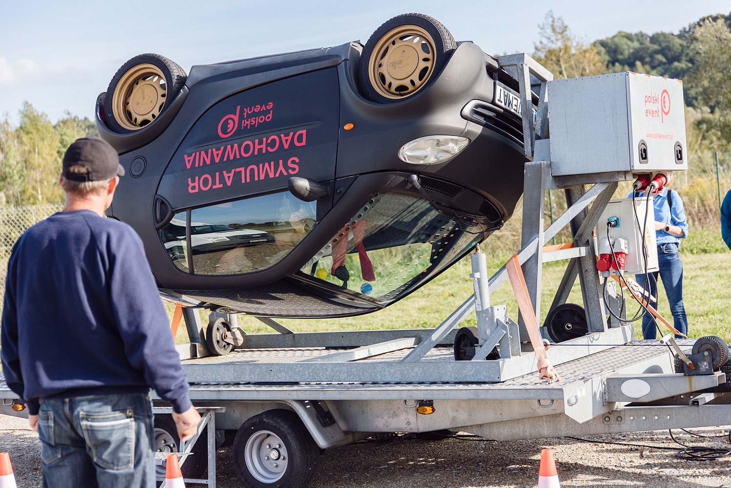 Czarny samochód Smart na platformie symulującej dachowanie, z widocznym logo firmy eventowej i napisem 'Symulator Dachowania'. Osoby obserwują demonstrację na zewnątrz.
