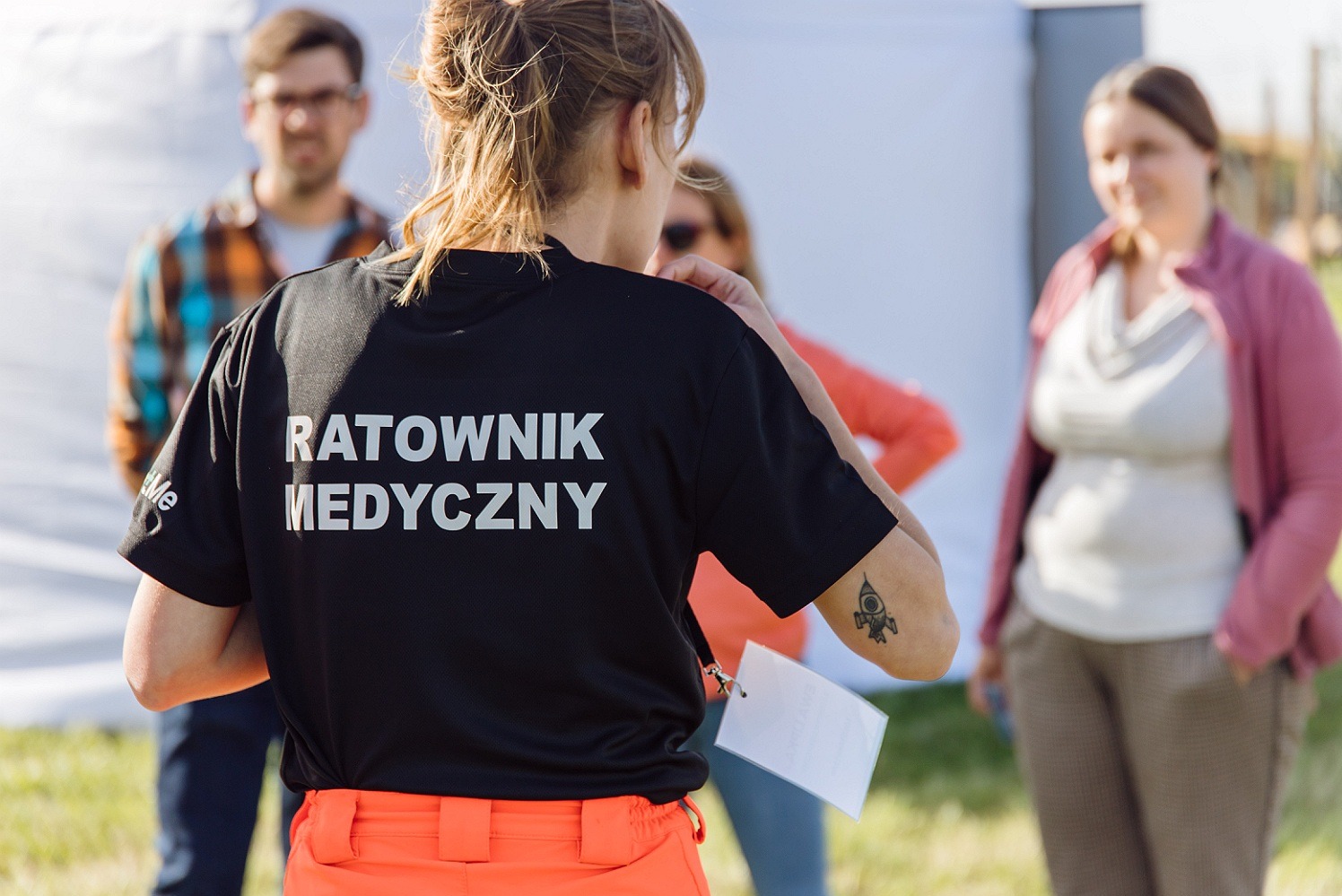Ratownik medyczny w czarnej koszulce z napisem na plecach, widoczny tatuaż rakiety na ramieniu, w tle uczestnicy szkolenia i biały namiot.