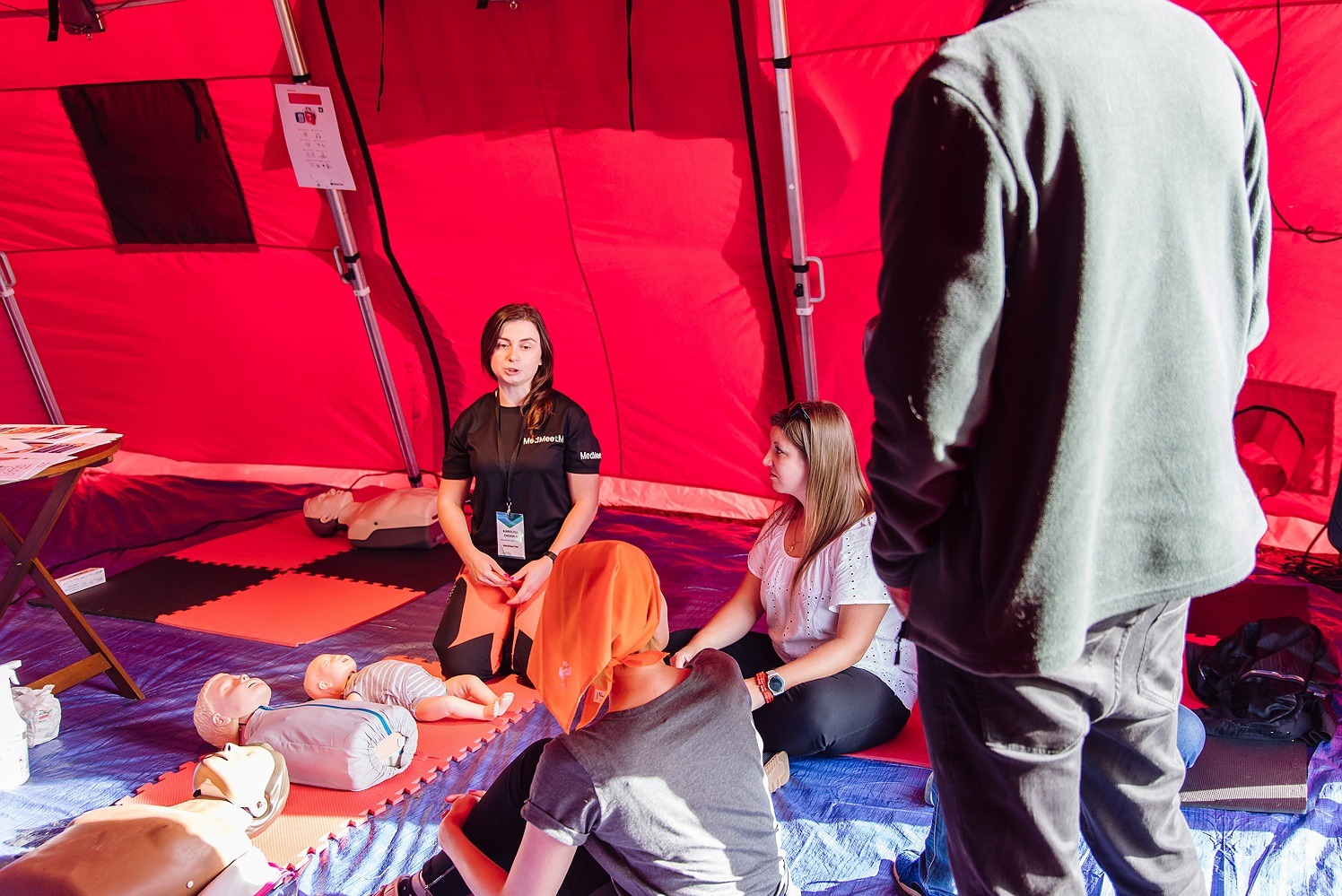 Wnętrze czerwonego namiotu, gdzie instruktorka w czarnej koszulce z logo firmy prezentuje techniki pierwszej pomocy na manekinach niemowląt i dorosłych. Uczestnicy siedzą na podłodze, słuchając...