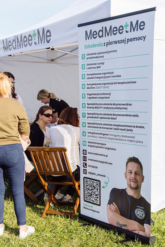 Stoisko promocyjne firmy MedMeetMe oferującej szkolenia z pierwszej pomocy, widoczne osoby siedzące przy stoliku i baner reklamowy z ofertą szkoleń, QR kodem i wizerunkiem założyciela firmy.