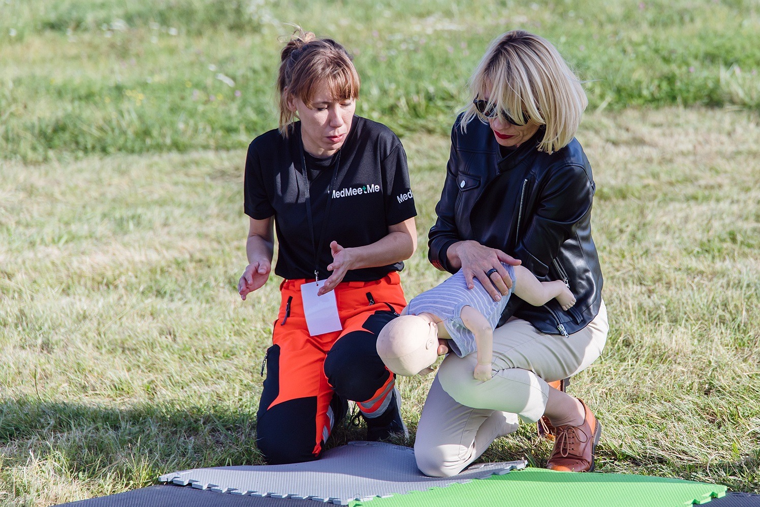 Instruktorka w czarnej koszulce z logo i pomarańczowych spodniach demonstruje na manekinie niemowlęcia technikę pierwszej pomocy, podczas gdy uczestniczka w skórzanej kurtce obserwuje.