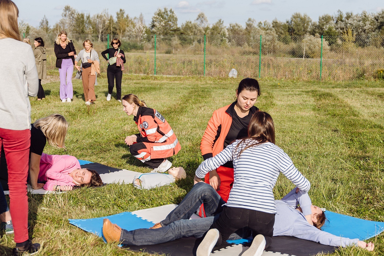 Uczestnicy kursu pierwszej pomocy ćwiczą resuscytację na fantomach na trawie podczas zajęć praktycznych na świeżym powietrzu, instruktor w pomarańczowej kurtce nadzoruje ćwiczenie.
