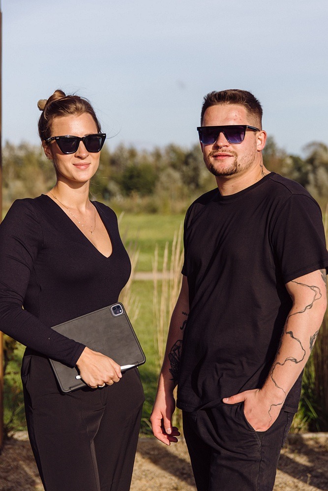 Dwoje ludzi w czarnych ubraniach i okularach przeciwsłonecznych, kobieta trzyma tablet, mężczyzna ma tatuaż na ramieniu, w tle zielona łąka.