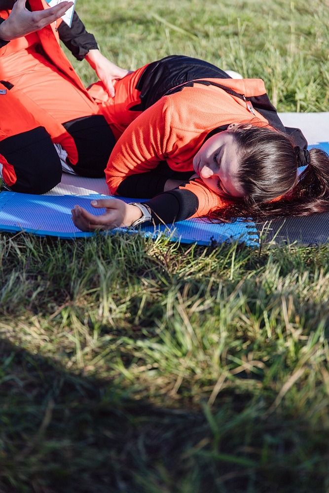 Kobieta w pomarańczowym stroju leży na niebieskiej macie na trawie podczas symulacji udzielania pierwszej pomocy. Instruktor sprawdza jej reakcję.