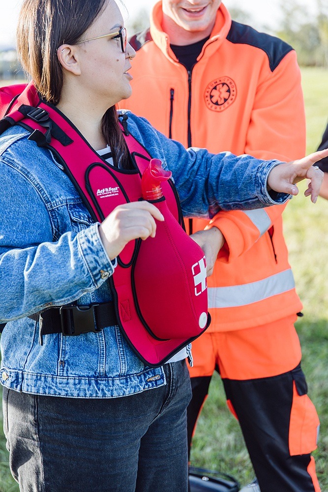 Kobieta w jeansowej kurtce i czerwonej kamizelce ratunkowej z ustnikiem do resuscytacji, obok mężczyzna w pomarańczowym uniformie medycznym z symbolem gwiazdy życia.
