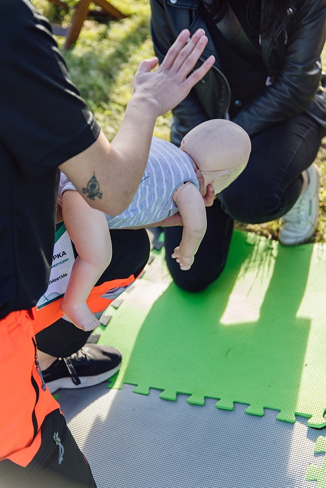 Instruktor z tatuażem na ramieniu demonstruje ułożenie manekina niemowlęcia podczas ćwiczeń resuscytacji na zielonej macie puzzlowej.