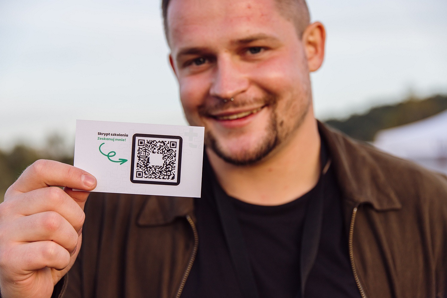 Uśmiechnięty mężczyzna z kolczykiem w nosie trzymający kartę z kodem QR i napisem 'Skrypt szkolenia. Zeskanuj mnie!'