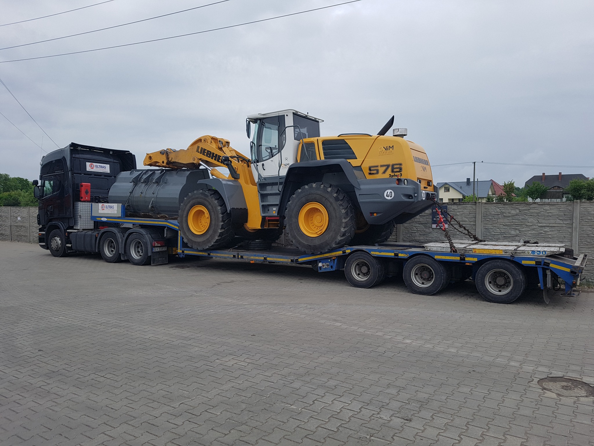 Ciężarówka z naczepą niskopodwoziową przewożąca żółto-białą ładowarkę kołową Liebherr 576, zabezpieczoną łańcuchami, na tle szarej kostki brukowej i ogrodzenia.