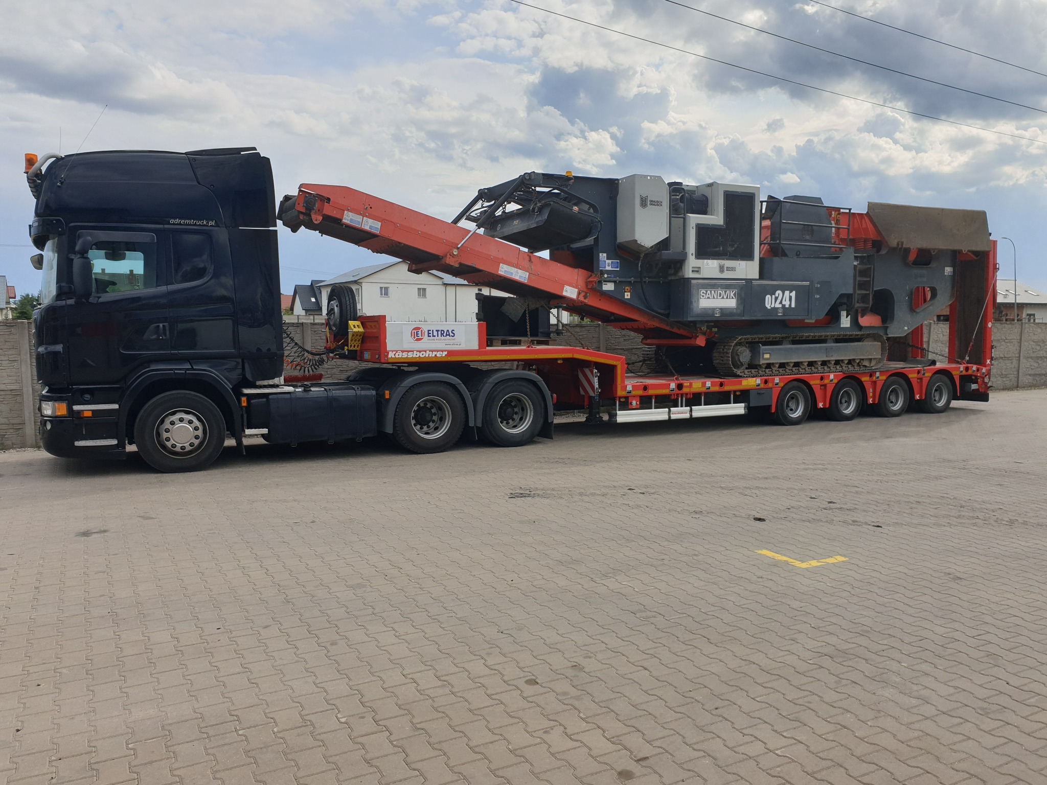 Czarny ciągnik siodłowy Scania z naczepą niskopodwoziową przewożącą mobilny przesiewacz Sandvik QJ241 z gąsienicowym podwoziem na brukowanym placu.