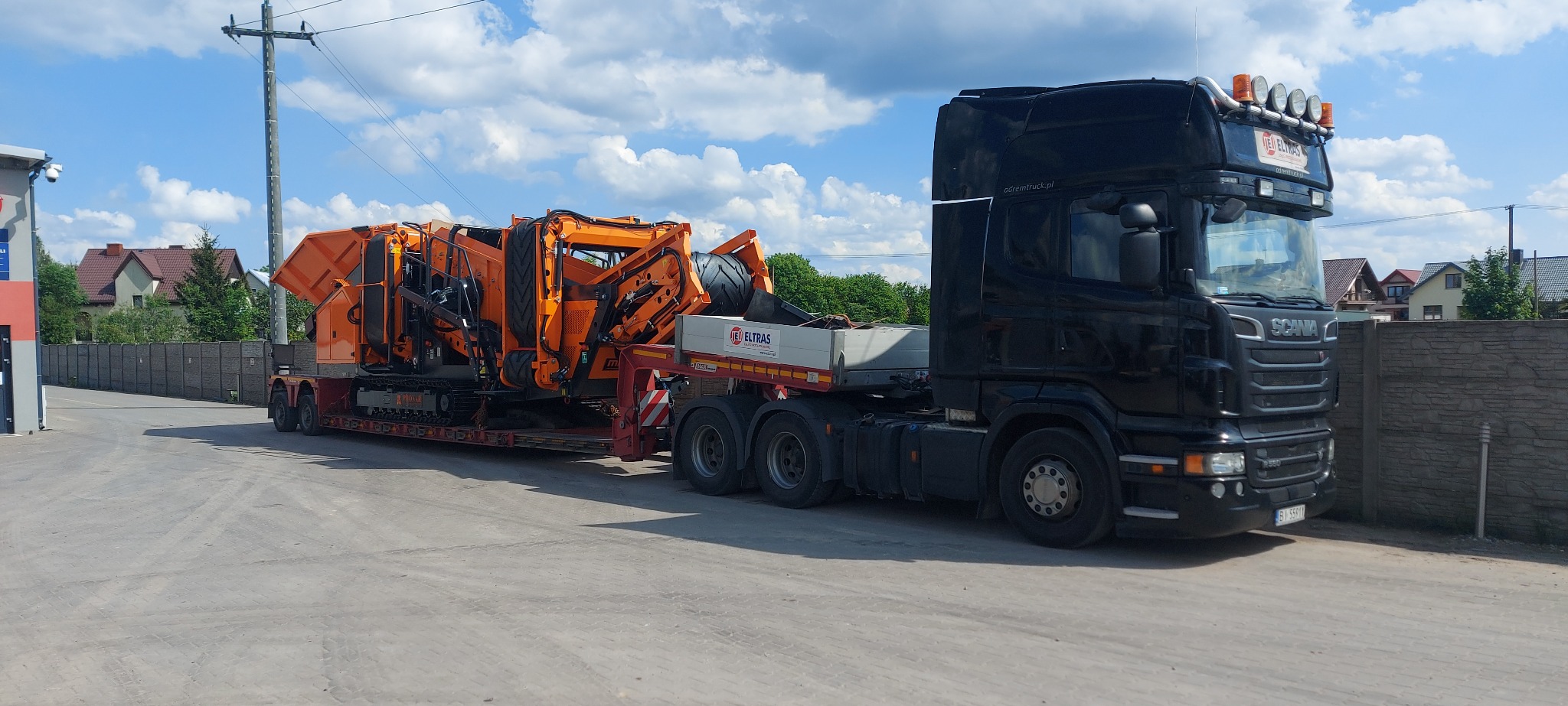 Czarna ciężarówka Scania z naczepą przewożąca pomarańczową maszynę do kruszenia kamieni, z logo firmy Eltas na naczepie, na tle błękitnego nieba z chmurami.