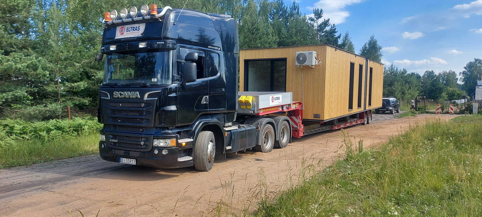 Czarna ciężarówka Scania przewozi modułowy dom z klimatyzacją na niskopodwoziowej naczepie, widok z boku na polnej drodze, drzewa w tle.