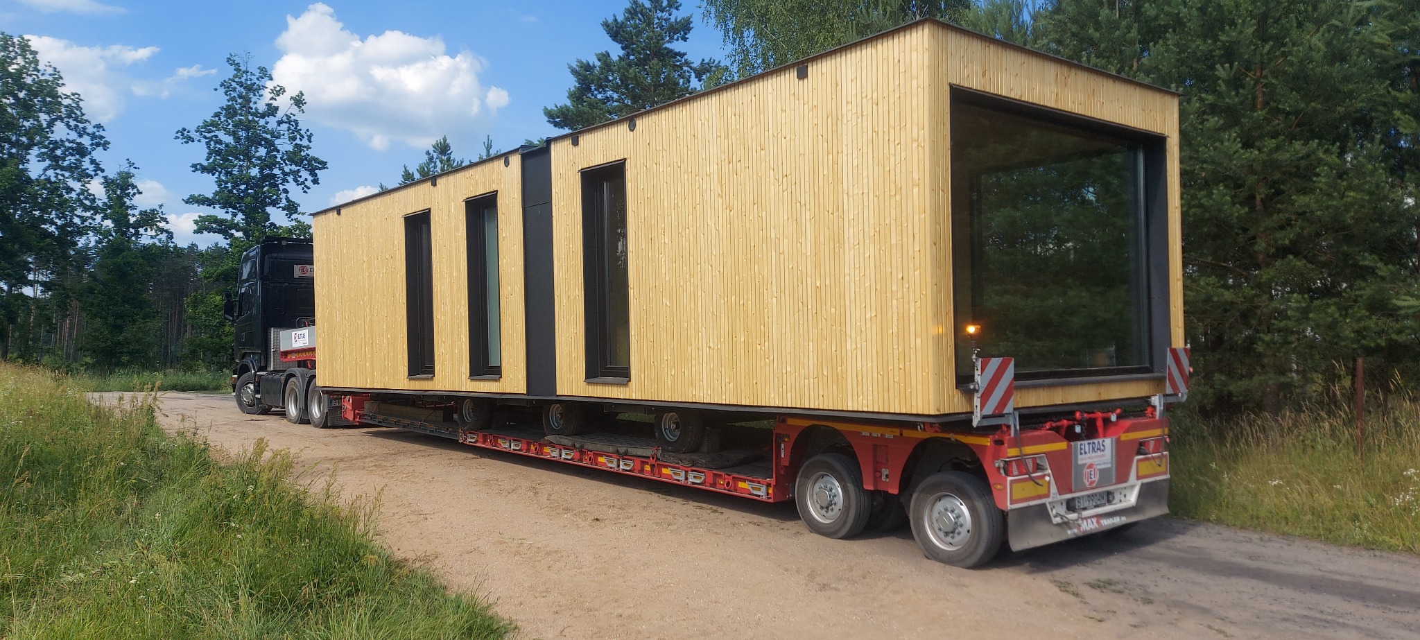 Ciężarówka przewozi modułowy, drewniany dom z dużym oknem, zabezpieczony na niskopodwoziowej platformie transportowej, na tle leśnej drogi.