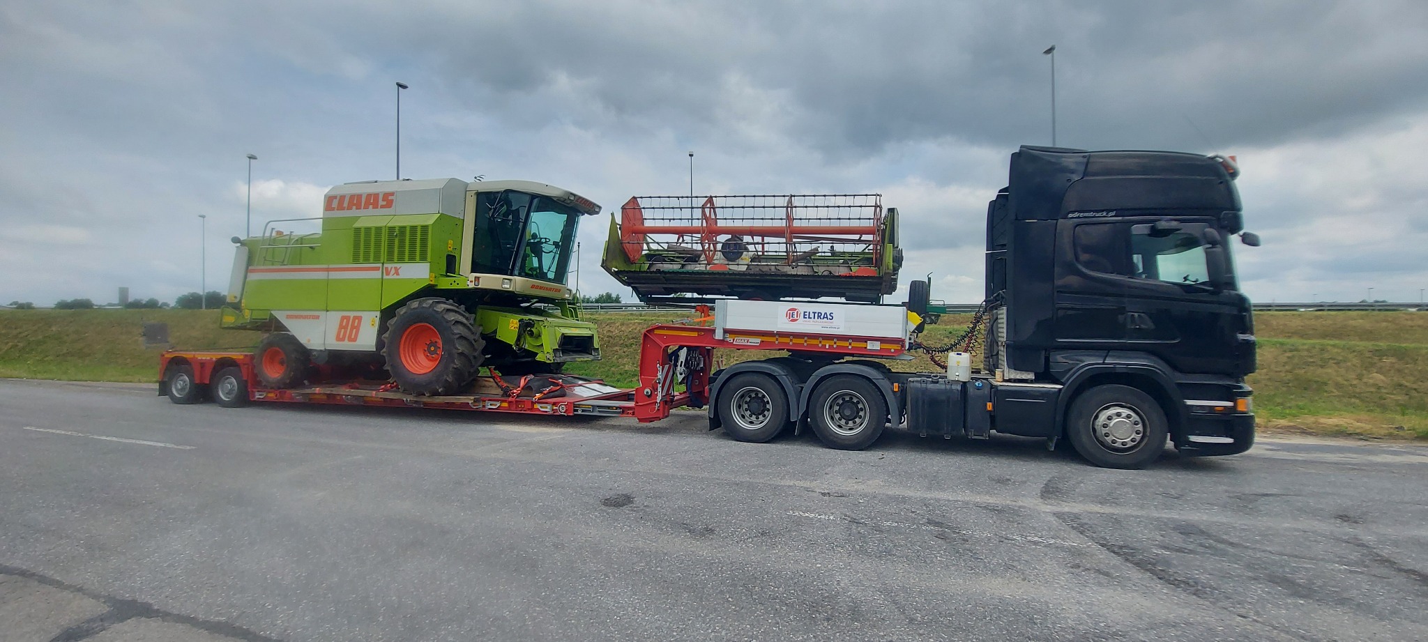 Czarny ciągnik siodłowy przewozi na niskopodwoziowej naczepie kombajn zbożowy Claas Dominator VX i jego heder na trasie. Widoczny logo firmy transportowej Eltraś.