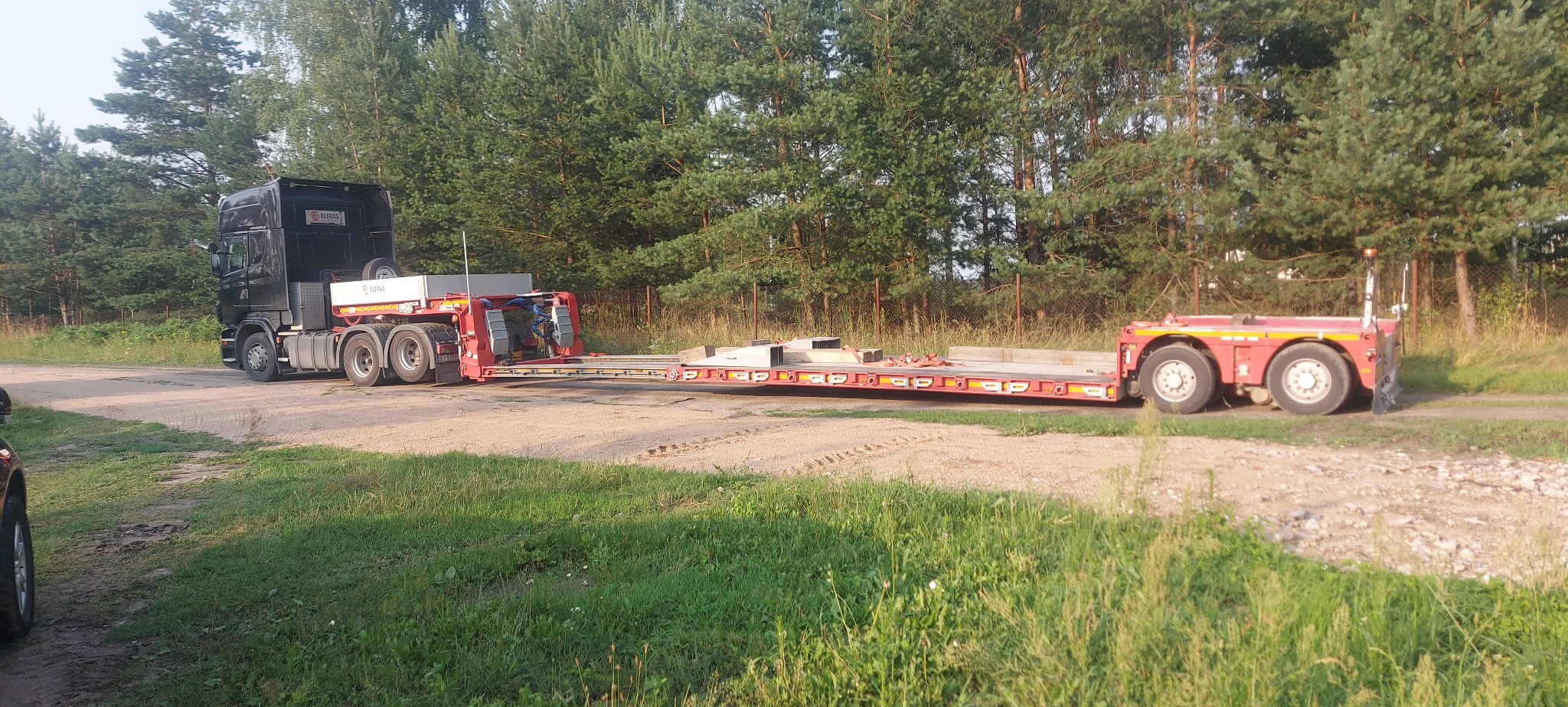 Czarny ciągnik siodłowy marki Scania z naczepą niskopodwoziową, przewożącą elementy konstrukcyjne na tle lasu.