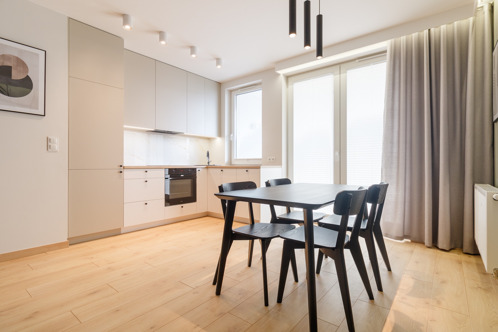 Nowoczesna kuchnia z jasnymi szafkami i drewnianą podłogą, czarny stół z krzesłami, minimalistyczne oświetlenie sufitowe, okno z żaluzjami i zasłonami.