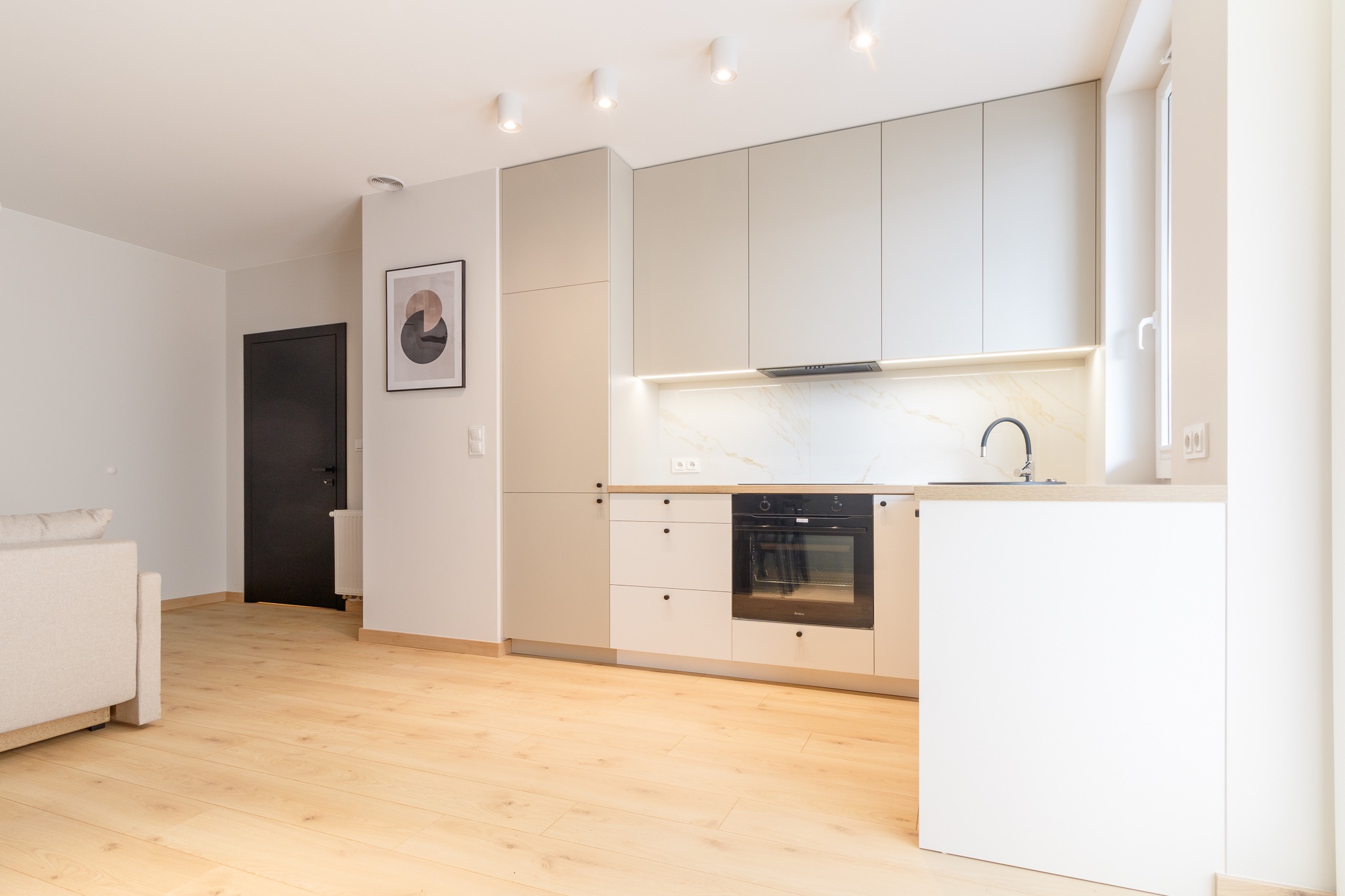 Jasna kuchnia w nowoczesnym stylu z drewnianą podłogą, białymi szafkami, marmurowym blatem i czarną armaturą, oświetlona punktowymi lampami sufitowymi i listwą LED pod szafkami górnymi.