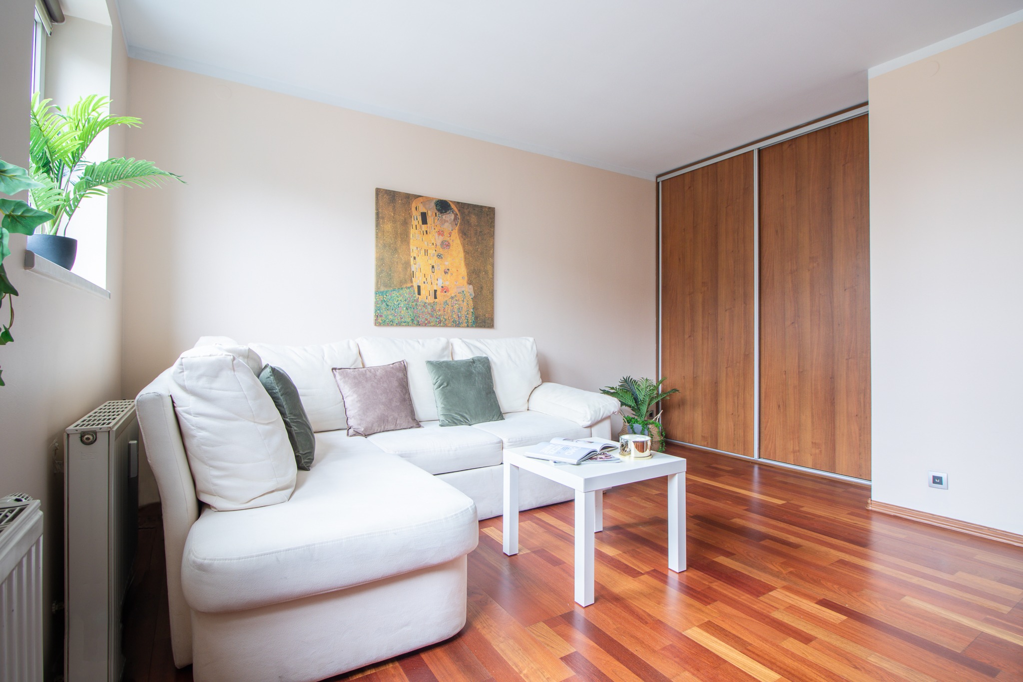 Jasny salon z białym narożnikiem, obrazem Klimta na ścianie, drewnianą podłogą i przesuwną szafą w tle.