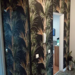 Fragment wnętrza z ciemną tapetą w motywy tropikalne, widoczne gniazdka elektryczne, fragment drzwi do innego pomieszczenia z widocznym akwarium i ceglaną ścianą.