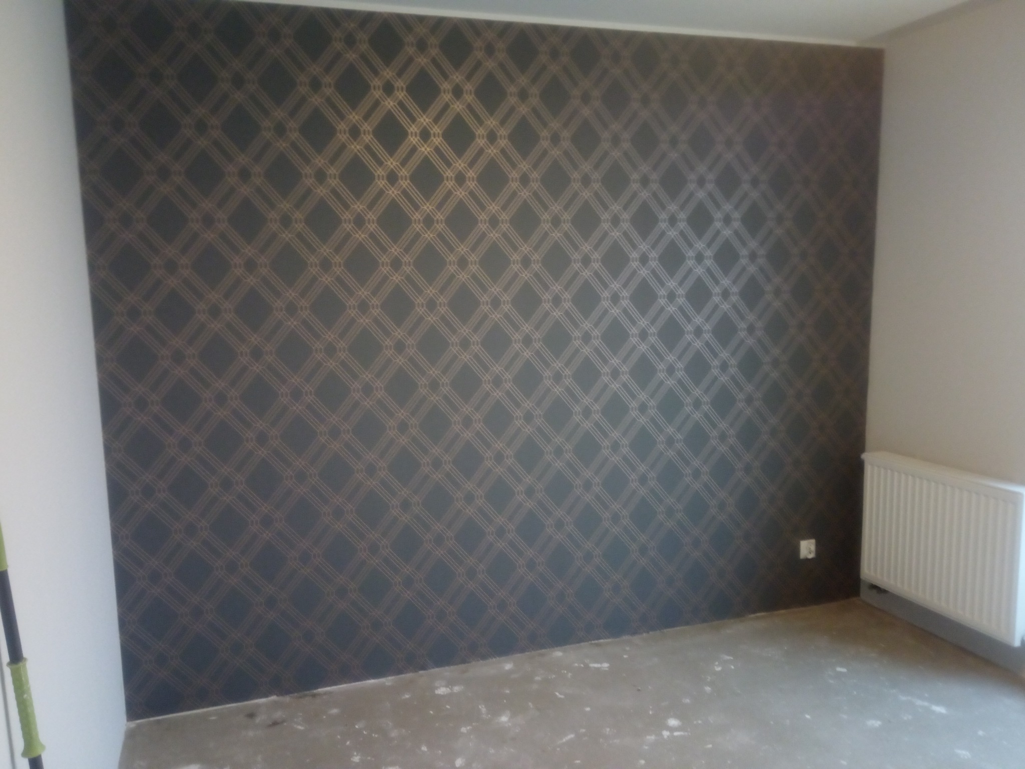 Ściana pokryta ciemną tapetą w geometryczny wzór z delikatnymi, złotymi liniami, widoczny fragment grzejnika i gniazdka elektrycznego, surowa podłoga w trakcie prac wykończeniowych.