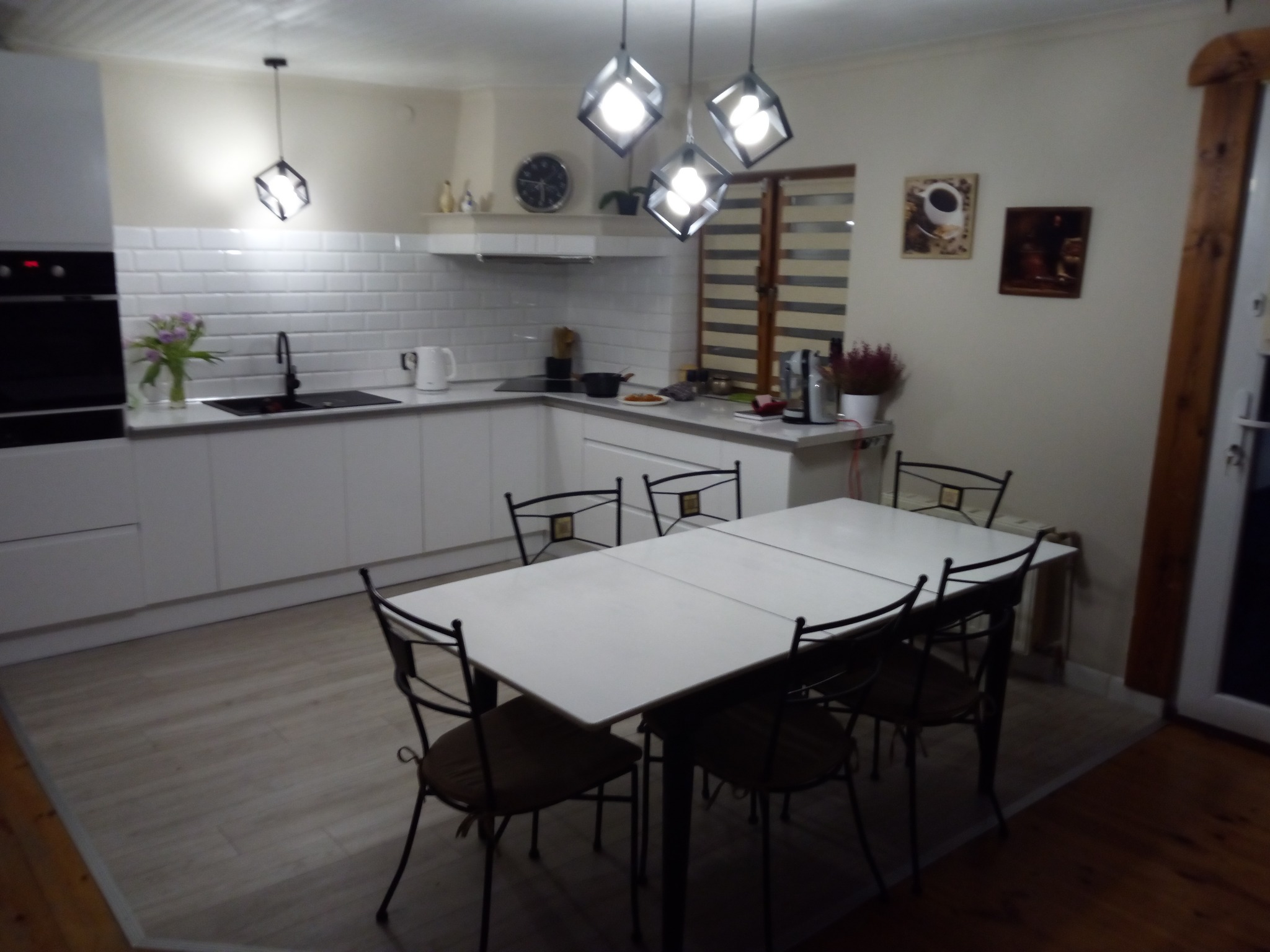 Jasna kuchnia z białymi szafkami, płytkami na ścianie i białym stołem z krzesłami, oświetlona wiszącymi lampami w kształcie kostek.