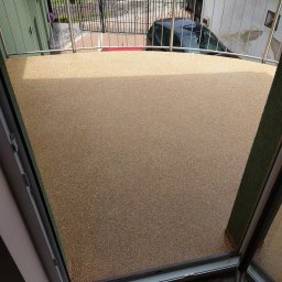 AM-system usługi budowlane - Balkon z nową, żywiczną posadzką w odcieniach beżu i brązu, widok z góry przez otwarte drzwi balkonowe, widoczne metalowe barierki balkonu i elementy otoczenia budynku.