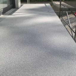 AM-system usługi budowlane - Szara, teksturowana posadzka balkonu z widocznymi rusztowaniami w tle, okno przesuwne z lewej strony kadru.