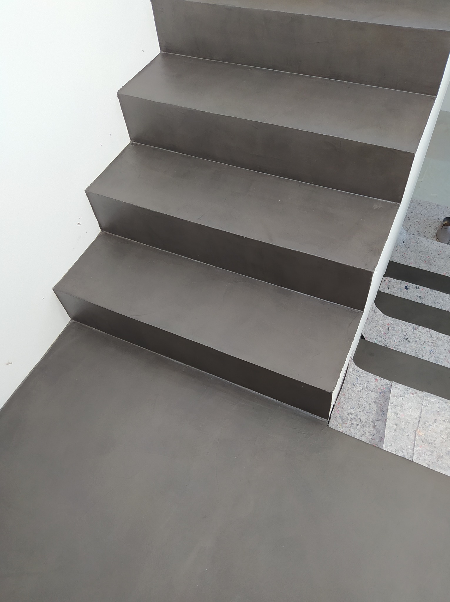 Schody pokryte jednolitą, szarą żywicą dekoracyjną NATURAL, widoczne z góry, z fragmentem zabezpieczonej podłogi w tle.