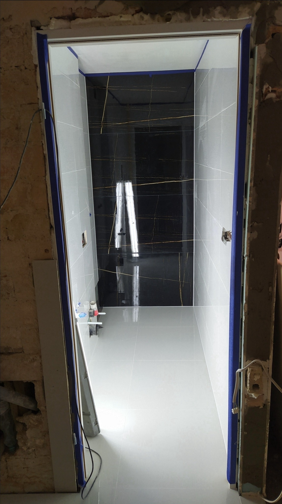 Wnętrze łazienki w trakcie remontu, widok przez ościeżnicę z nieotynkowanymi ścianami, białe płytki podłogowe i szare ścienne, z czarnym pasem płytek z żyłkami złota pod sufitem.