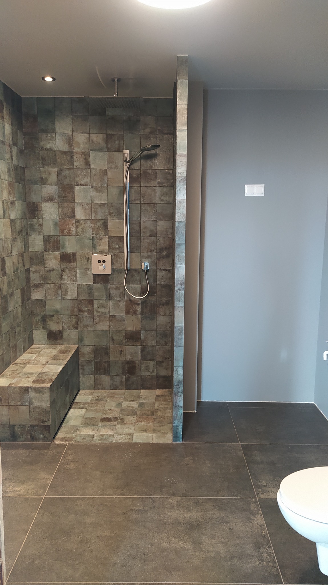 Nowoczesna kabina prysznicowa z siedziskiem wyłożona mozaiką w odcieniach brązu i zieleni, widoczny panel sterowania i deszczownica, na podłodze duże, ciemne płytki.