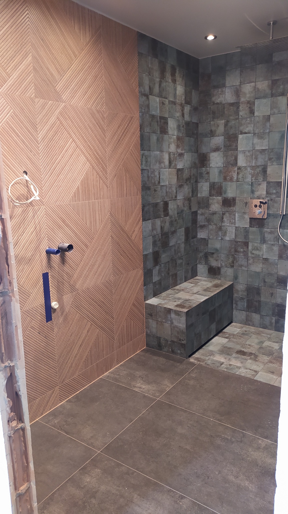 Wnętrze łazienki w trakcie remontu, widoczne płytki ścienne w dwóch wzorach: geometryczny brązowy i kwadratowy szary, oraz szare płytki podłogowe. Na ścianie widoczne wystające rury instalacji...