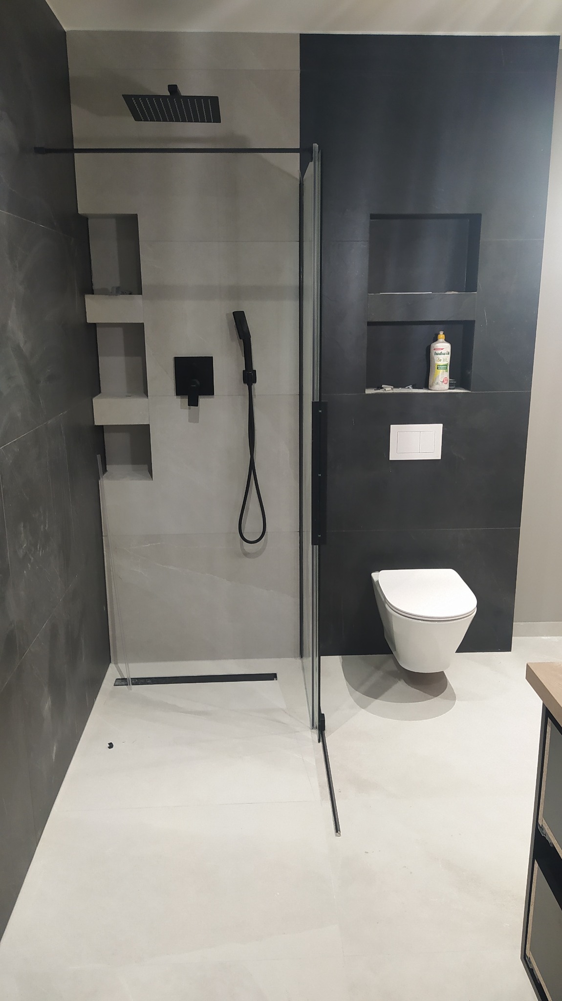 Nowoczesna łazienka z kabiną prysznicową typu walk-in, czarną armaturą i wnękami w ścianie wykończona szaro-czarnymi płytkami.