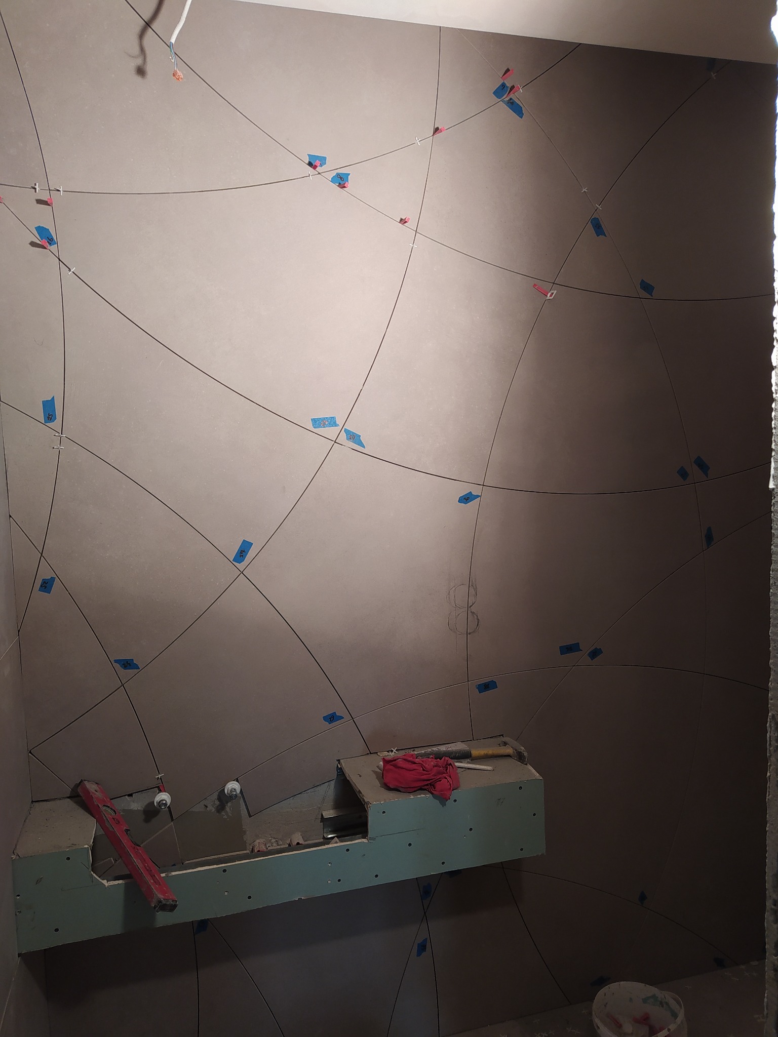 Ściana łazienki w trakcie układania płytek z zaznaczonym wzorem, użyte niebieskie taśmy i krzyżyki dystansowe, widoczny fragment zabudowy pod umywalkę i narzędzia.