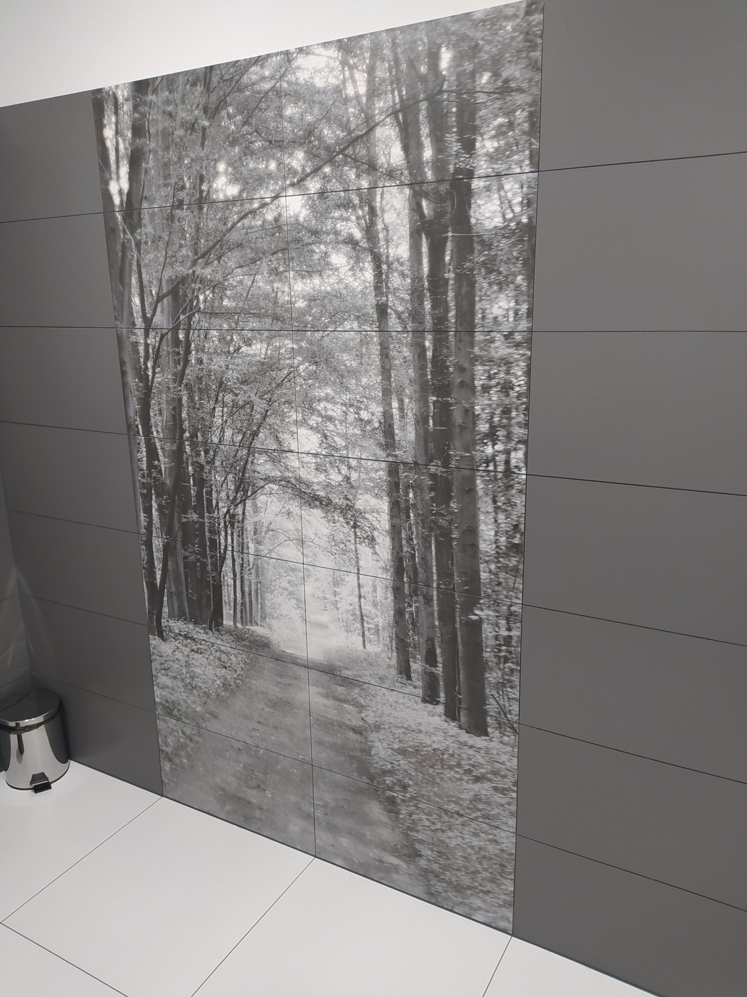 Ściana łazienki z szarymi płytkami imitującymi drewno i dużym panelem z nadrukiem czarno-białego leśnego krajobrazu, biała podłoga i chromowany kosz na śmieci w rogu.